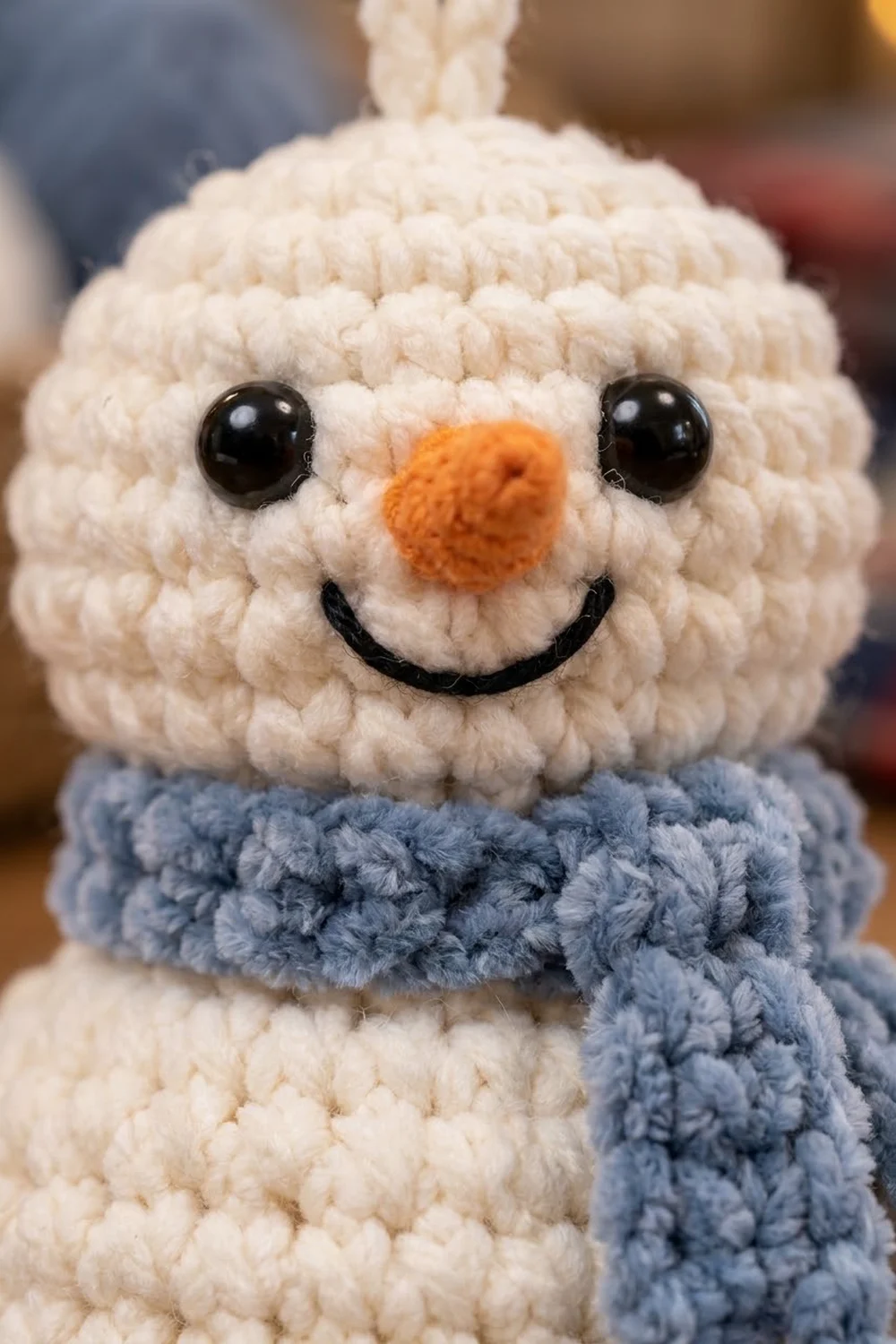 Crocheted snowman with textured cream body, black eyes, orange nose, embroidered smile, and blue scarf.