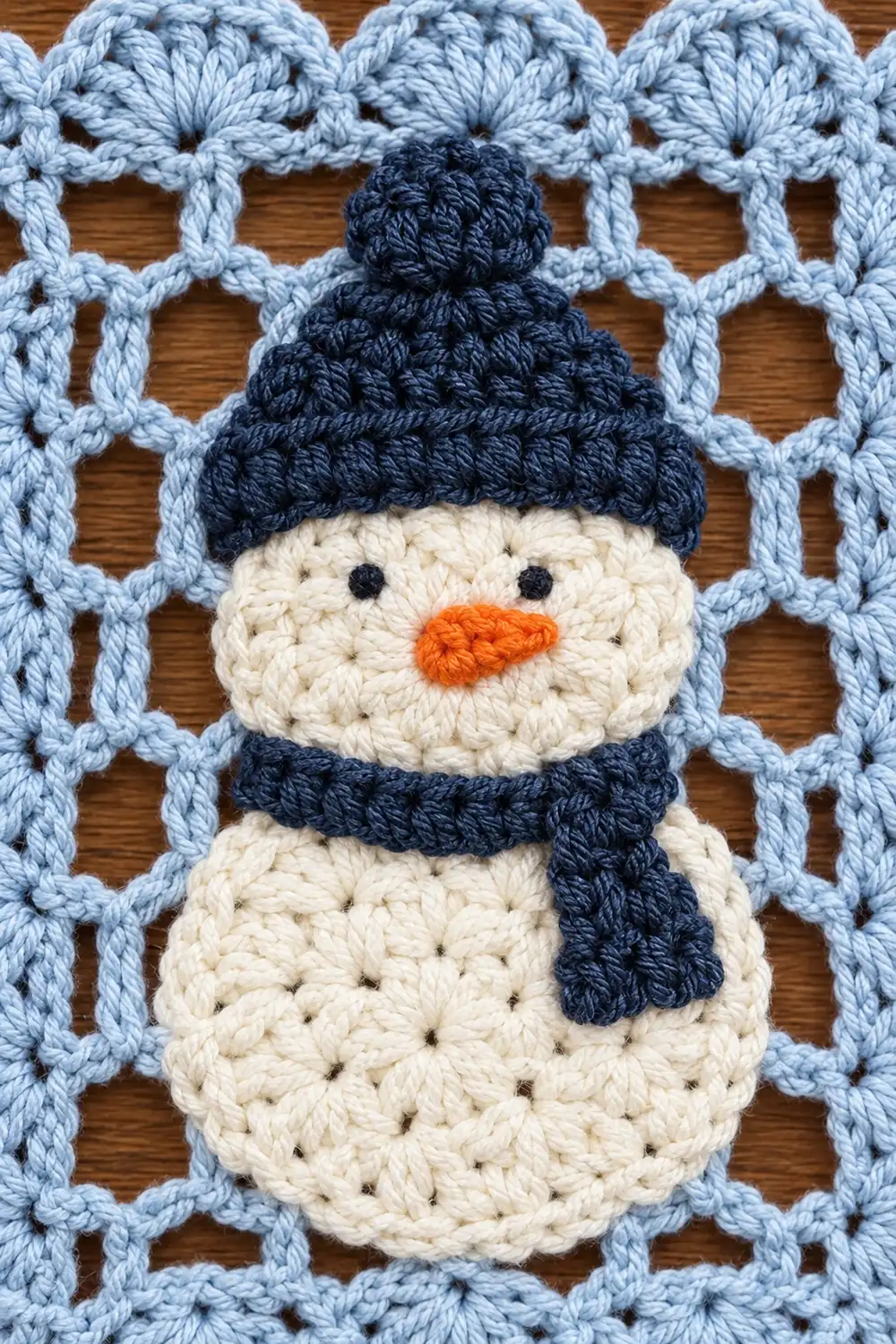 A crocheted snowman with cream body, navy hat and scarf, and orange carrot nose on a light blue lattice background.