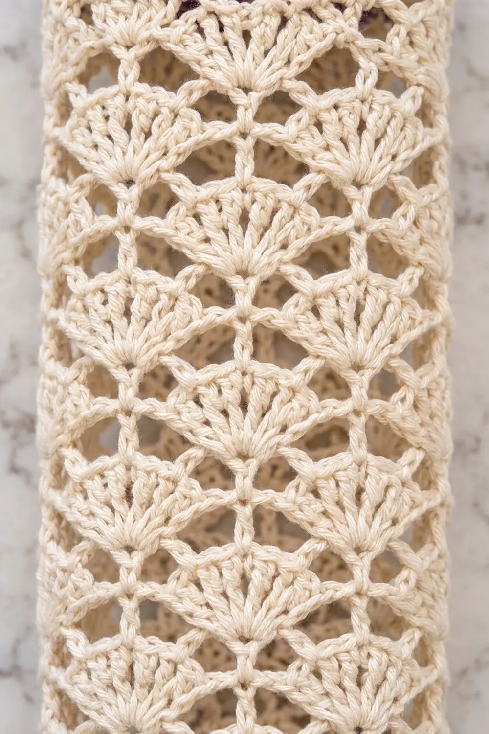 Cream-colored crochet lace with repeating openwork fan motifs, showing delicate thread texture against a soft, blurred background.
