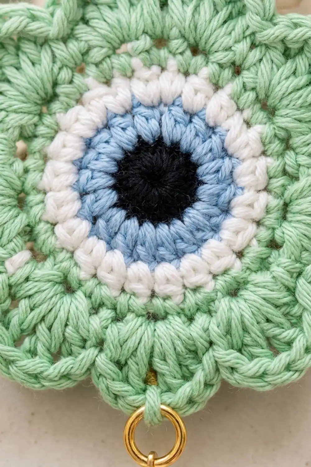 Crochet eye motif with concentric rings of light green, white, blue, and black yarn with gold metal ring attachment.