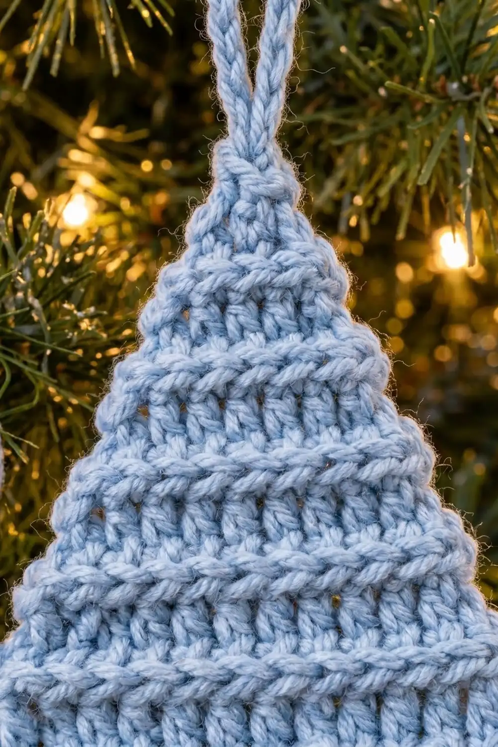 Light blue crocheted Christmas tree ornament hanging amid evergreen branches with warm golden bokeh lights.