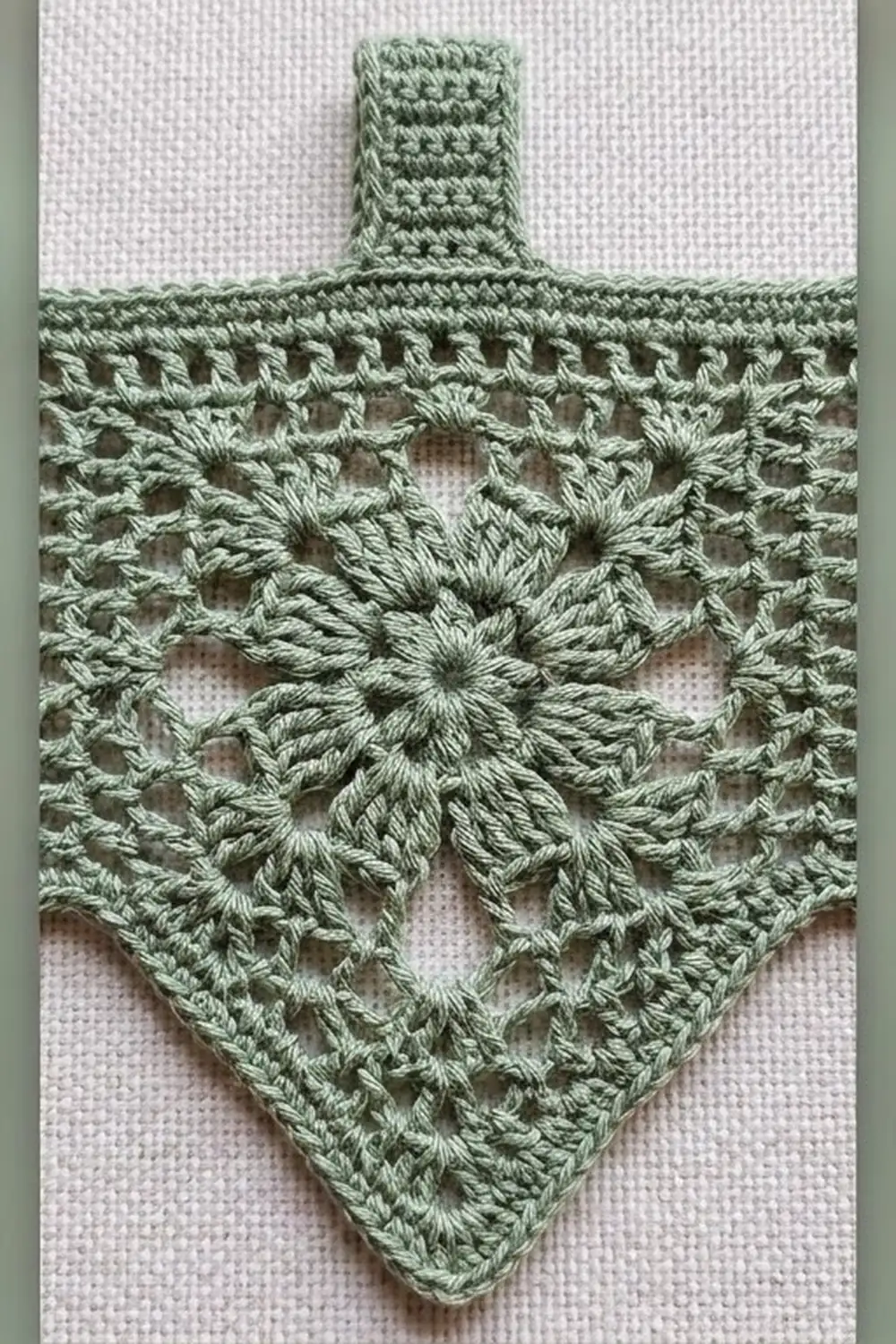 Sage-green crocheted cross ornament with openwork lace pattern and flower motif on white fabric background.