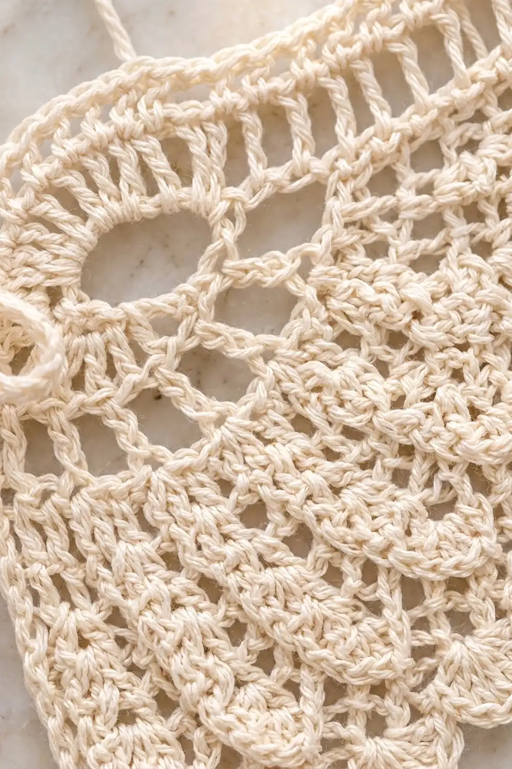Cream crocheted lace fabric with an intricate openwork pattern showcasing twisted yarn texture against a soft neutral background.