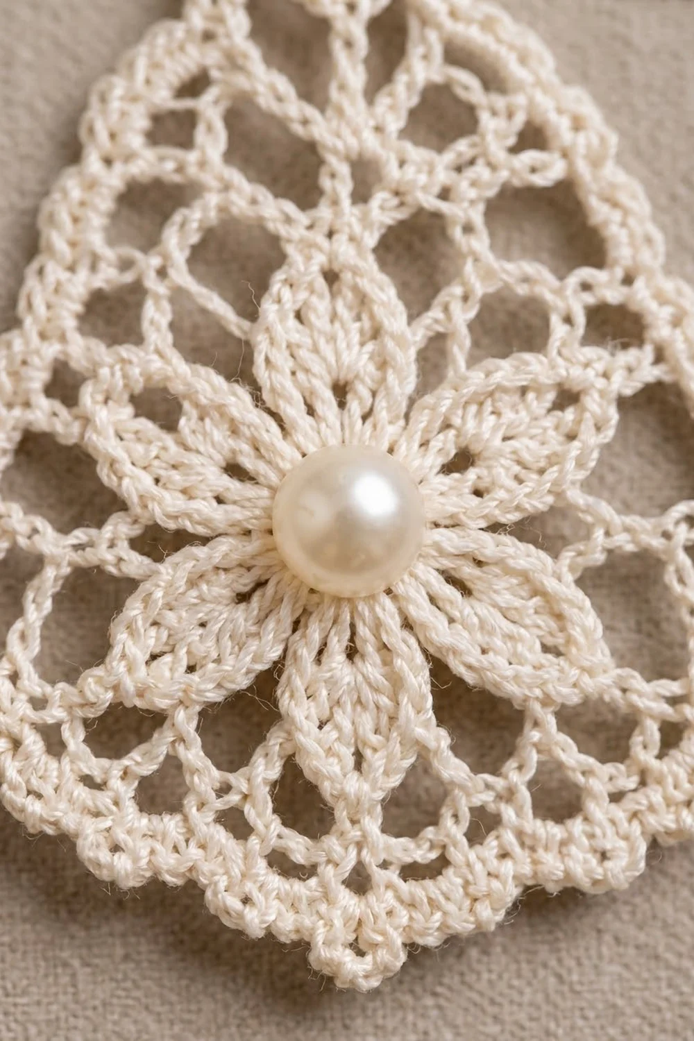 Cream-colored crochet flower with openwork lace petals centered with a pearl button on neutral beige fabric.
