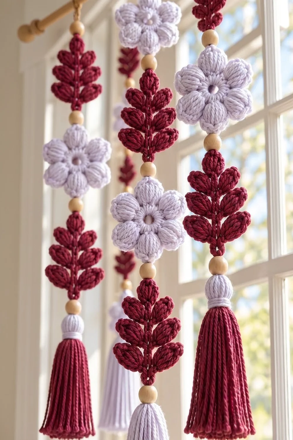 Crocheted floral garland with lavender flowers and burgundy leaves, beads, and tassels hanging in sunlit window.