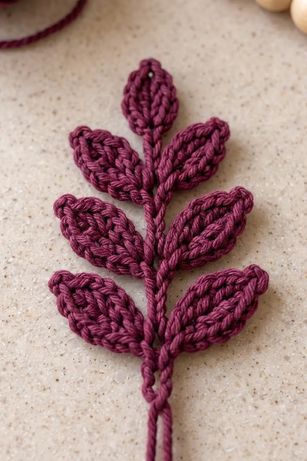 Burgundy crocheted plant with braided leaves arranged on a stem, set against a speckled cream surface.