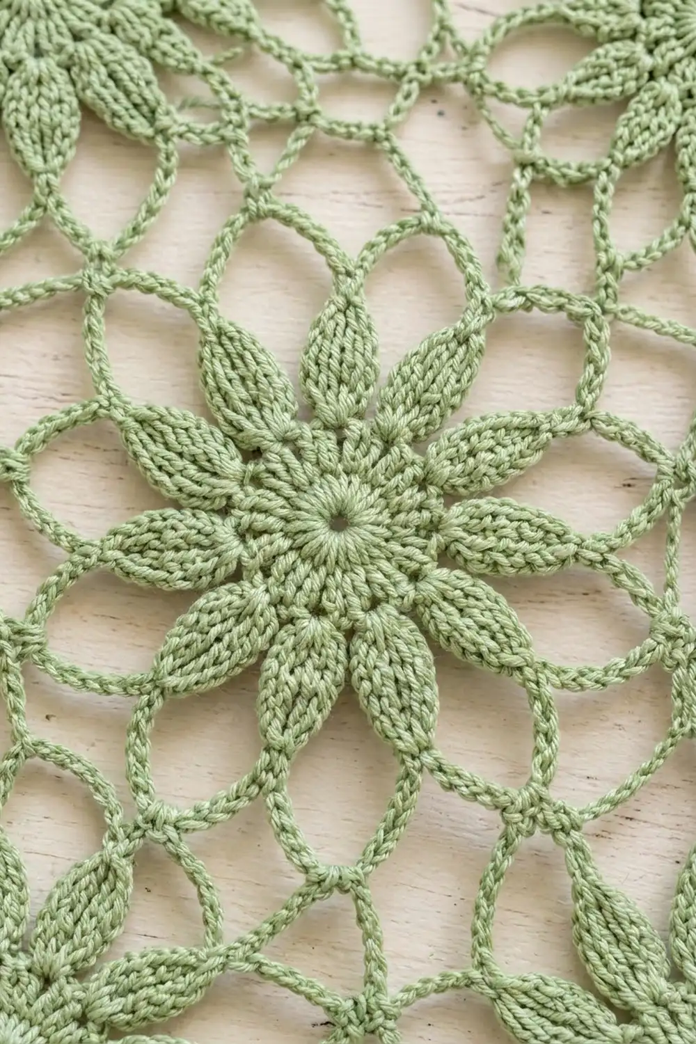 Sage-green crocheted flower with pointed petals and geometric mesh pattern on wooden surface.