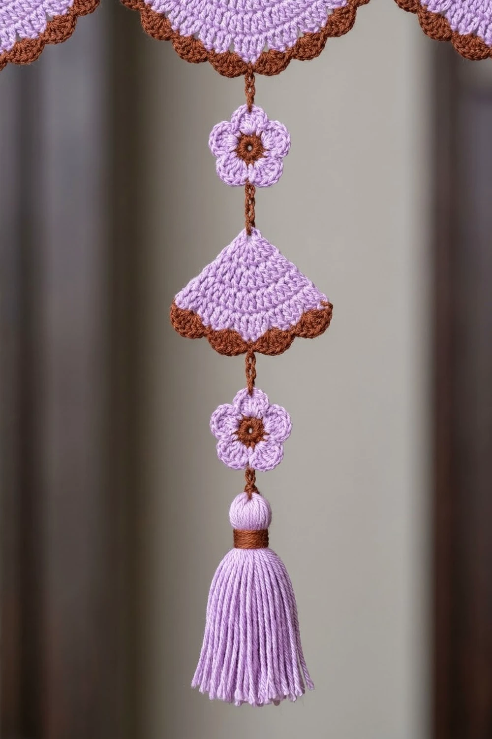 Crocheted purple and brown hanging ornament featuring flowers, bell center, and tassel against neutral gray background.