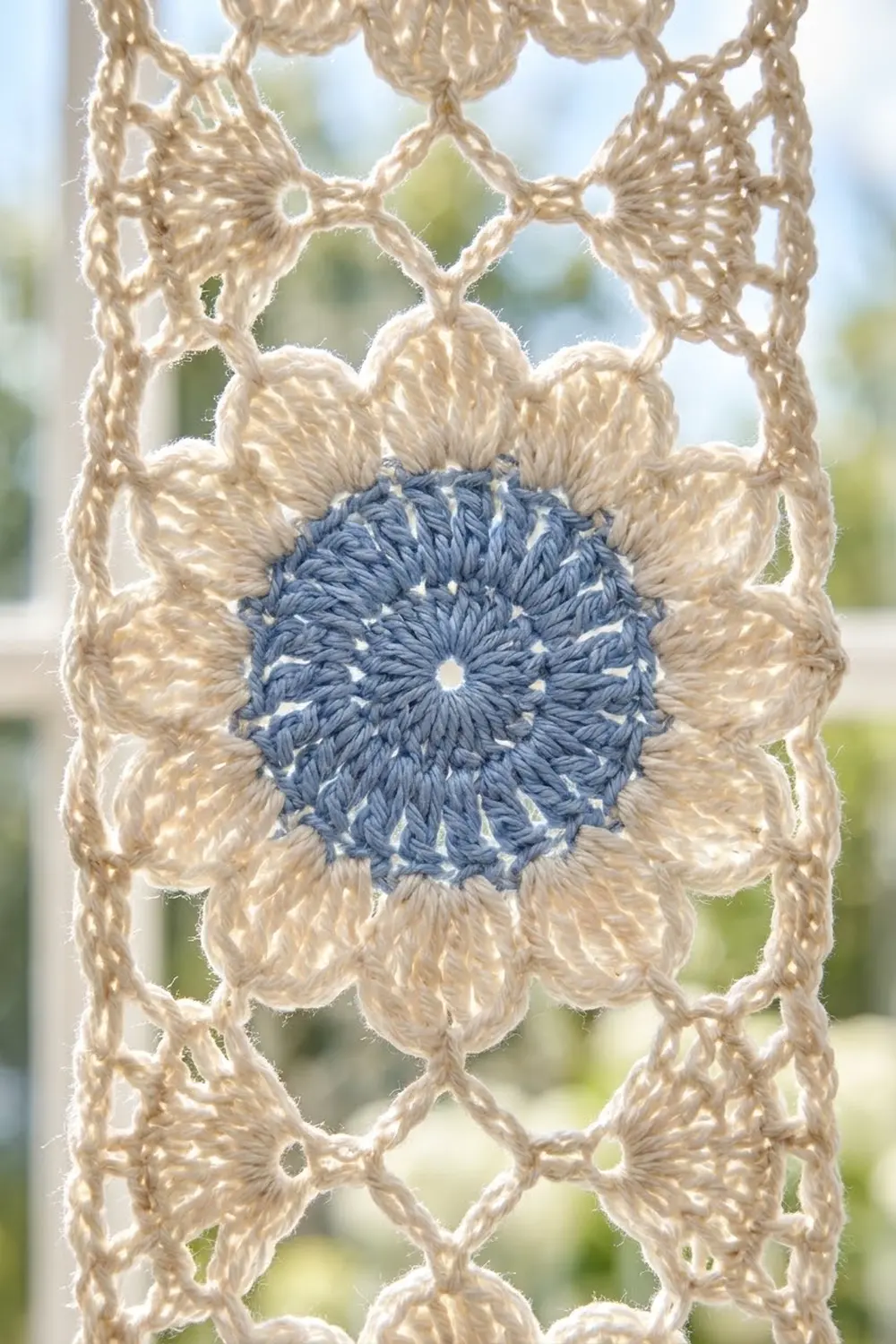 Cream-colored crocheted lace with geometric pattern features a blue flower motif, photographed outdoors with soft green background blur.