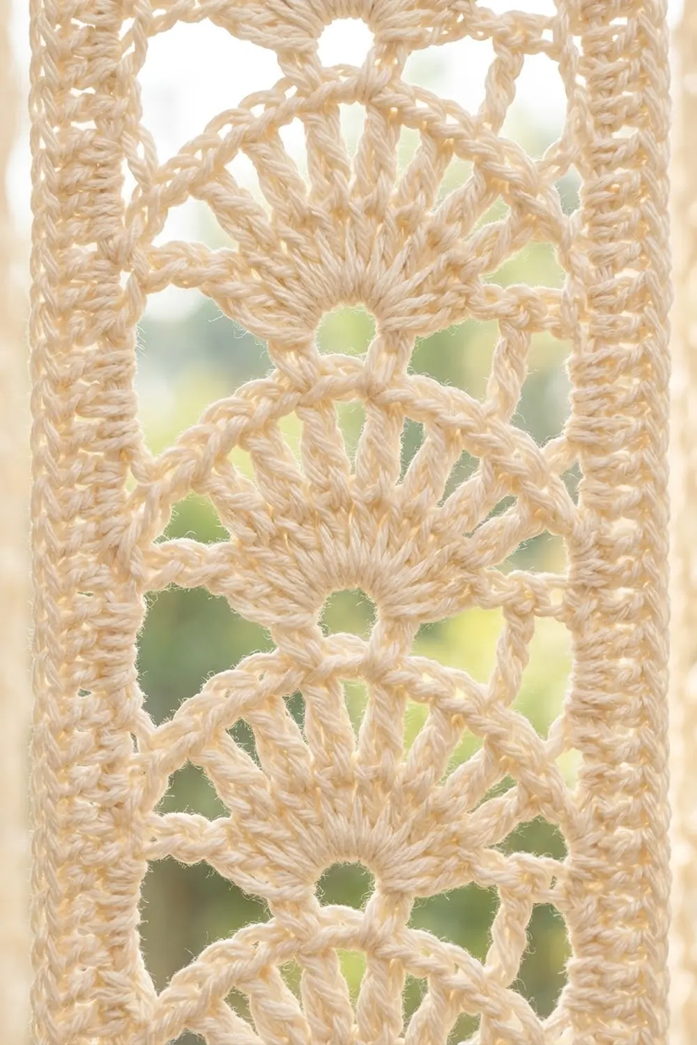 Close-up of natural-colored crocheted lace with geometric flower motifs and circular openings, backlit against blurred green foliage.