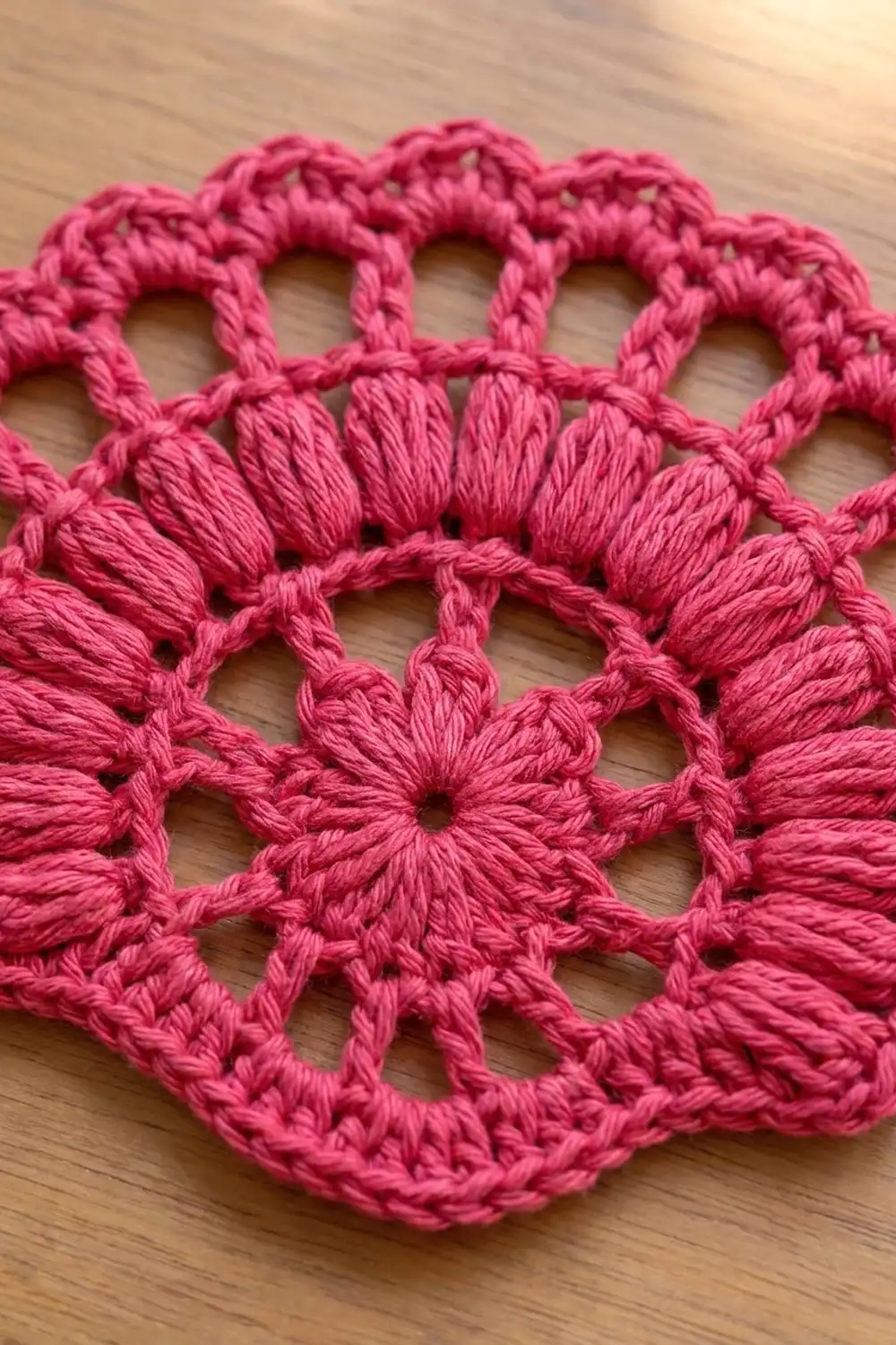 Bright pink crocheted doily with open floral motifs and intricate yarn texture detail on a wooden surface.