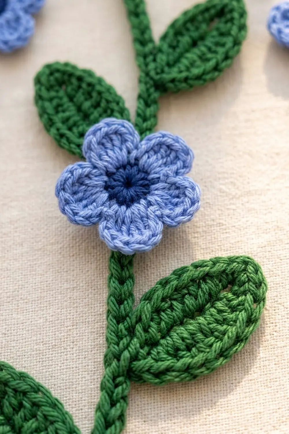 Light blue crocheted flower with five petals and dark center, green leaves, on cream linen fabric.