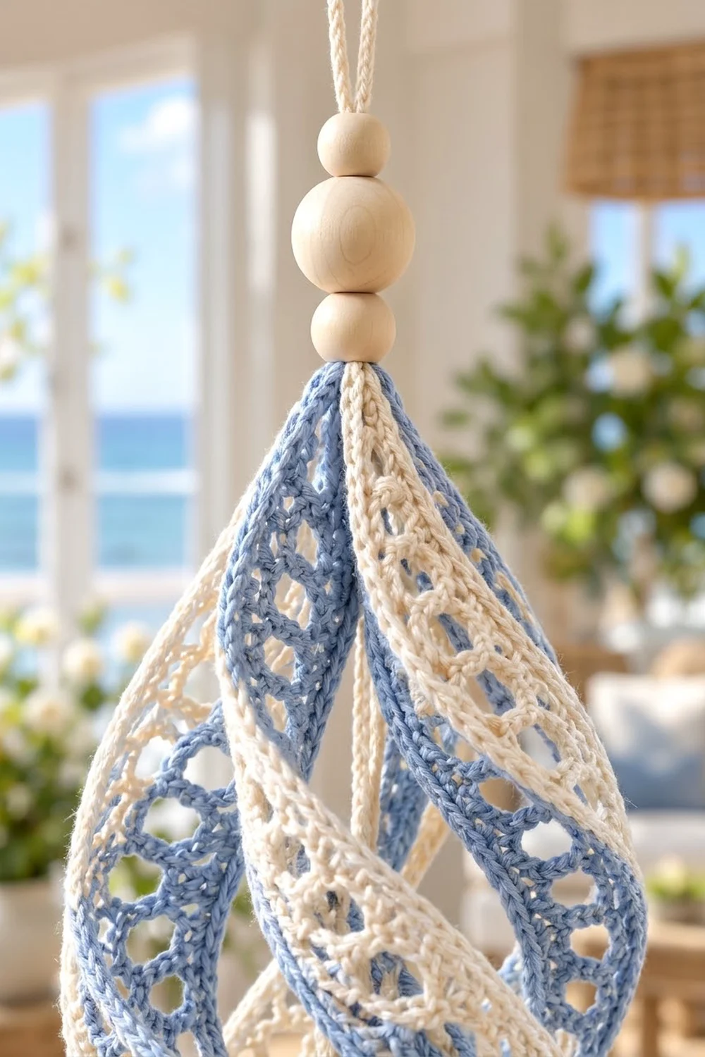 Blue and white crocheted teardrop ornament with stacked wooden beads hangs in sunny coastal room.