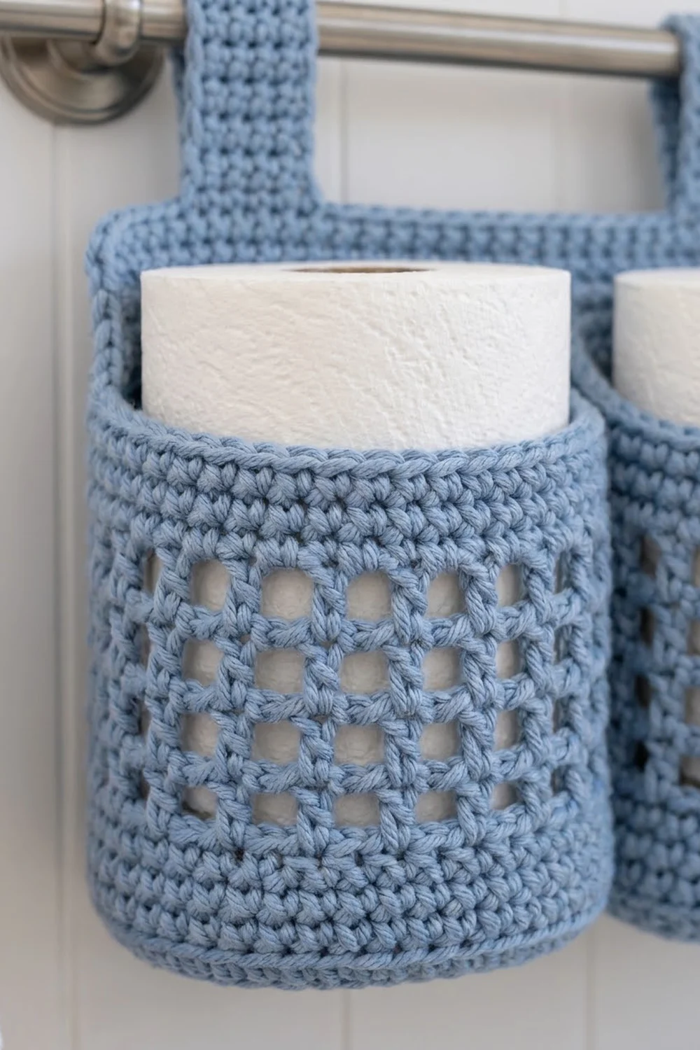 Light blue crocheted toilet roll holder with open-mesh pattern hanging from bathroom curtain rod.