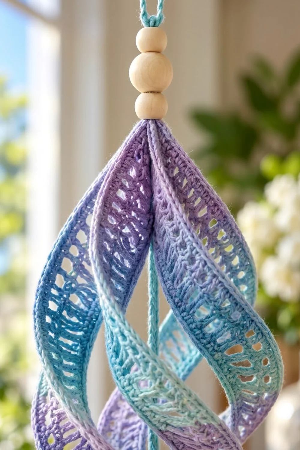 Crocheted lace ornament in purple-to-teal gradient with stacked wooden beads, hanging indoors near plants.