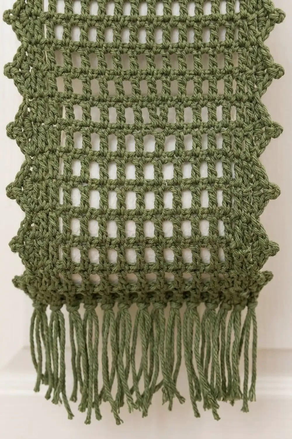 Sage green crocheted textile with open lattice stitch and hanging fringe trim on neutral backdrop.
