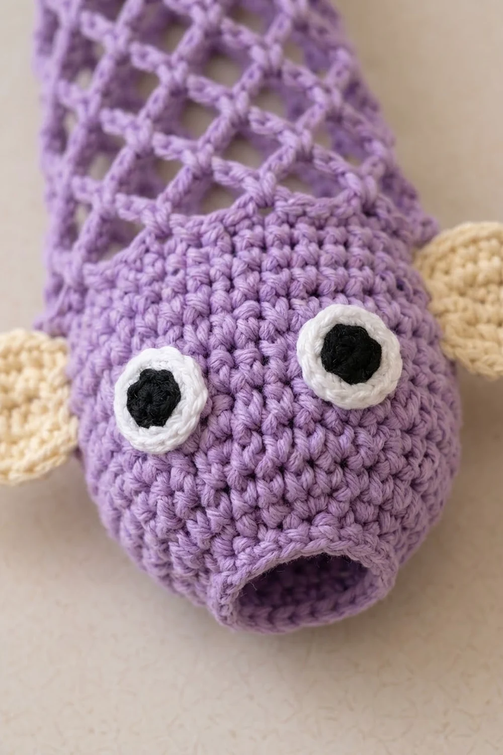 Close-up of a lavender crocheted bunny with white and black eyes, surrounded by tan-colored crocheted ears against a beige background.
