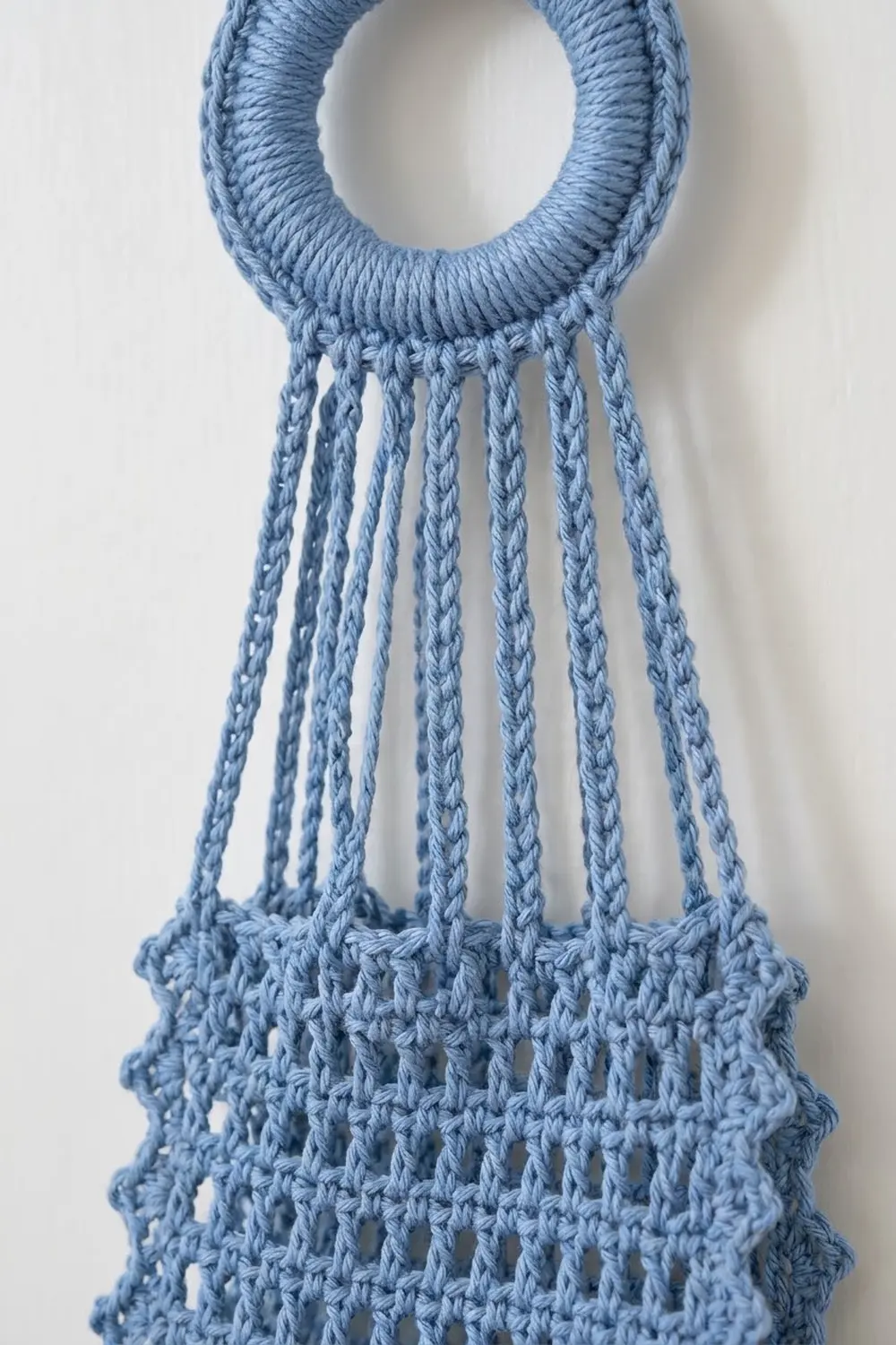 Dusty blue crocheted macramé plant hanger with wrapped handle and knotted yarn strands hanging down.