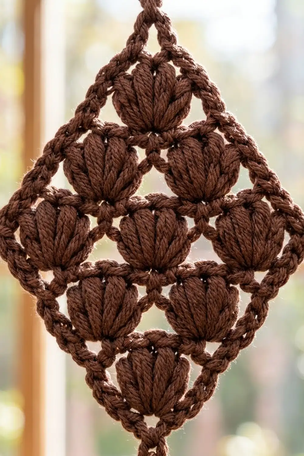 Brown crocheted lace ornament with textured puff stitch pattern arranged in a diamond shape against a softly blurred background.