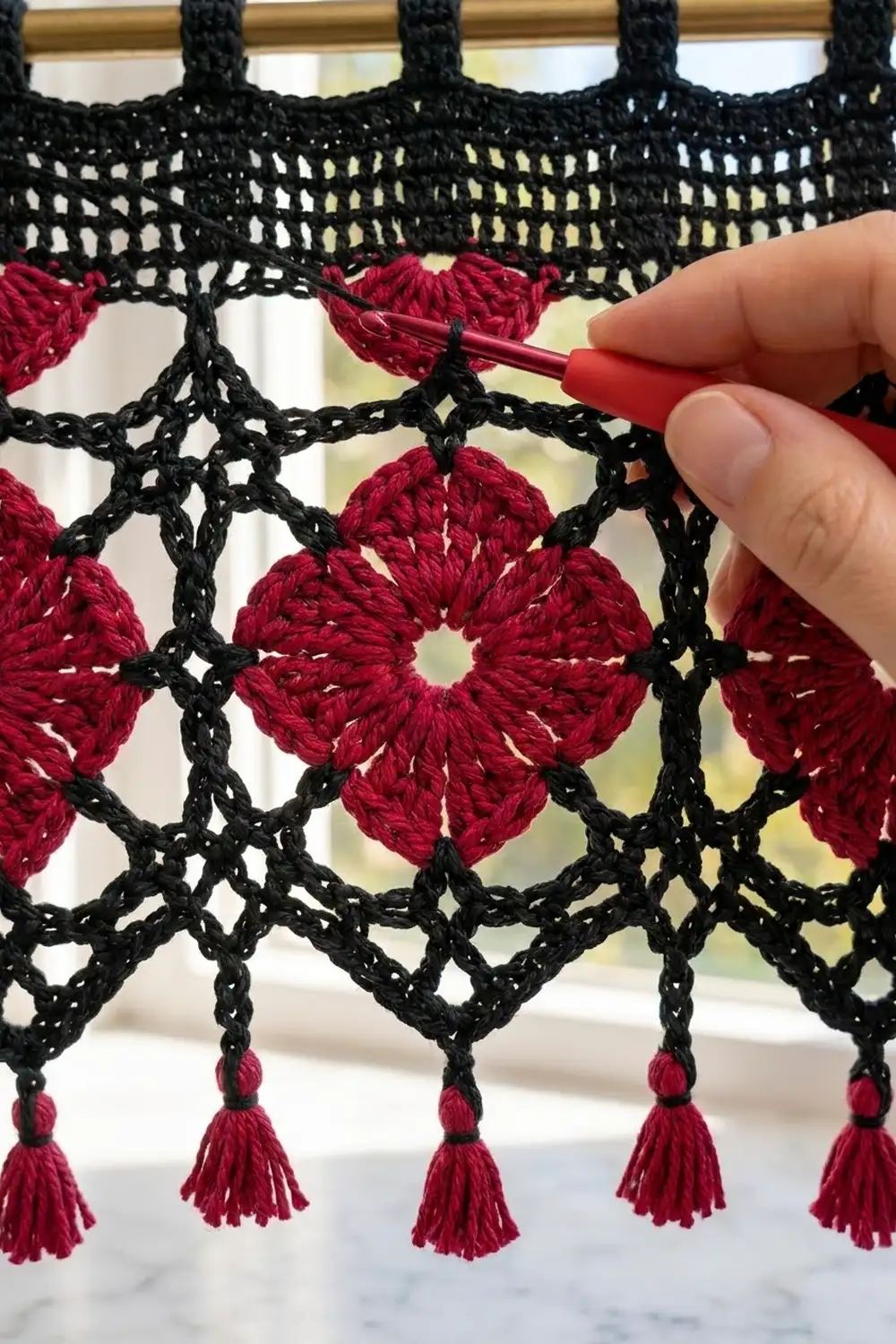 Black crochet lace with vibrant red rosettes and tassels being worked on with a crochet hook.