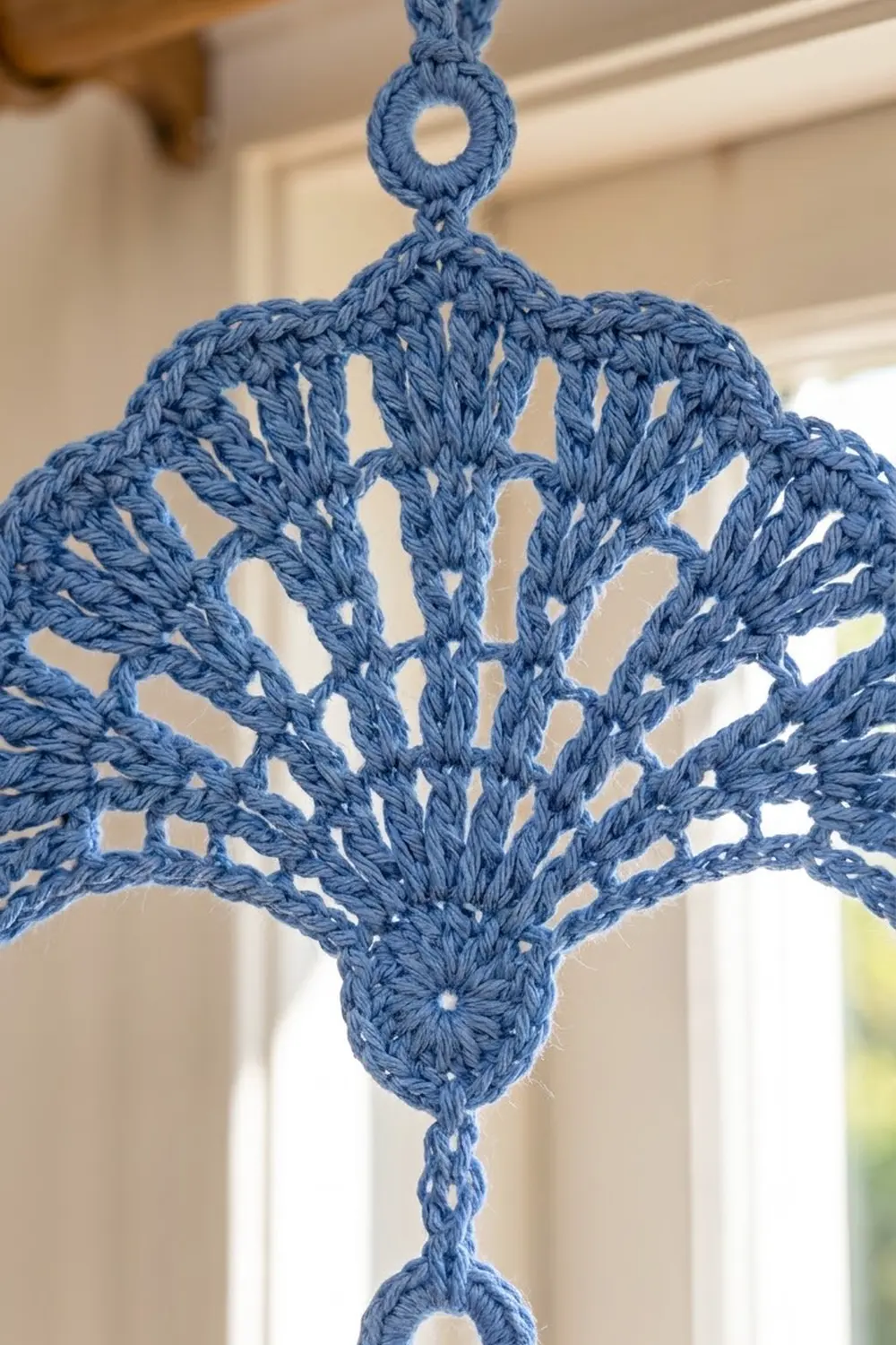 Blue crocheted star ornament with openwork diamond pattern hanging indoors against a blurred beige and white background.