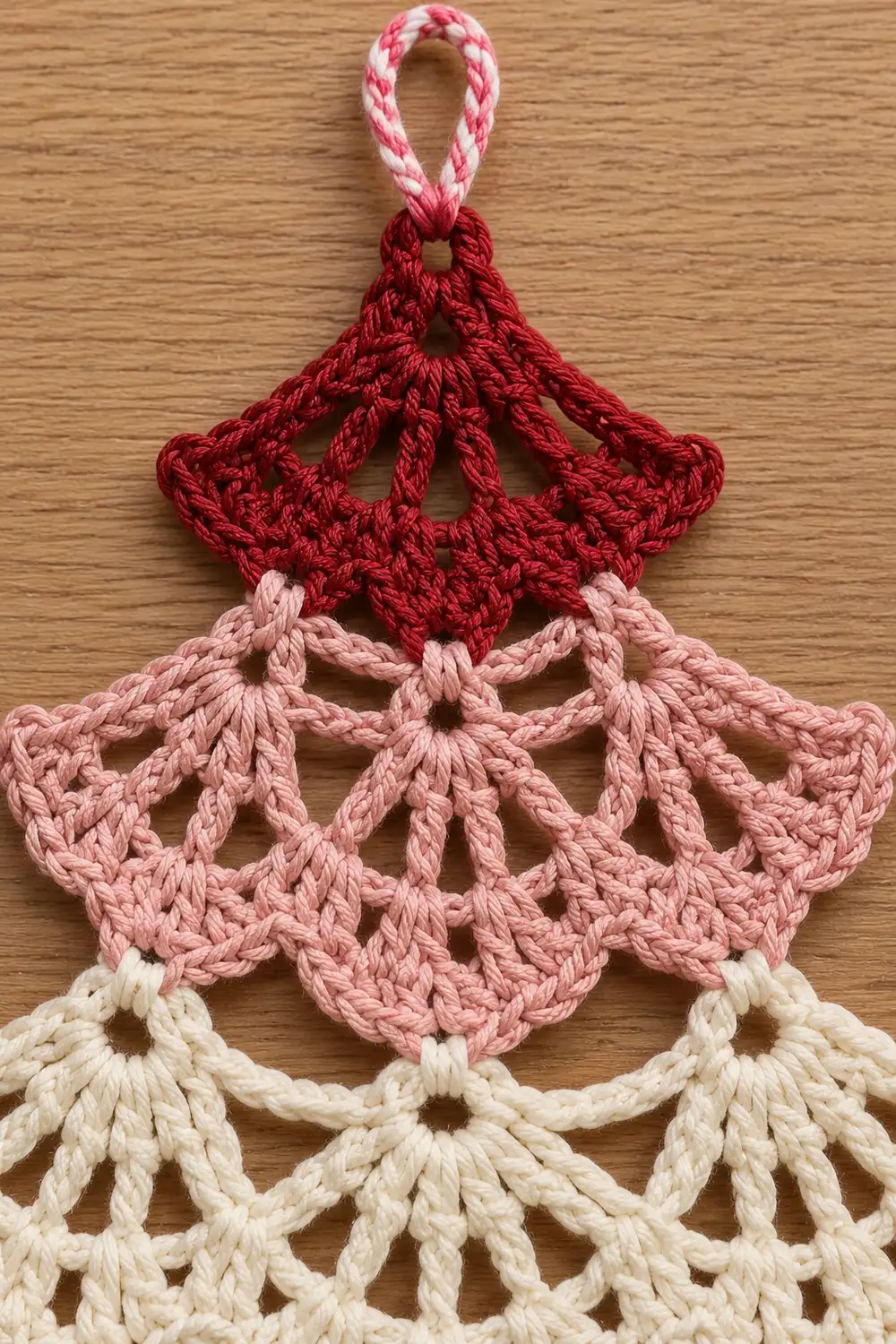 Crocheted ornament with red, pink, and cream layers hanging from a striped pink and white loop on wood.