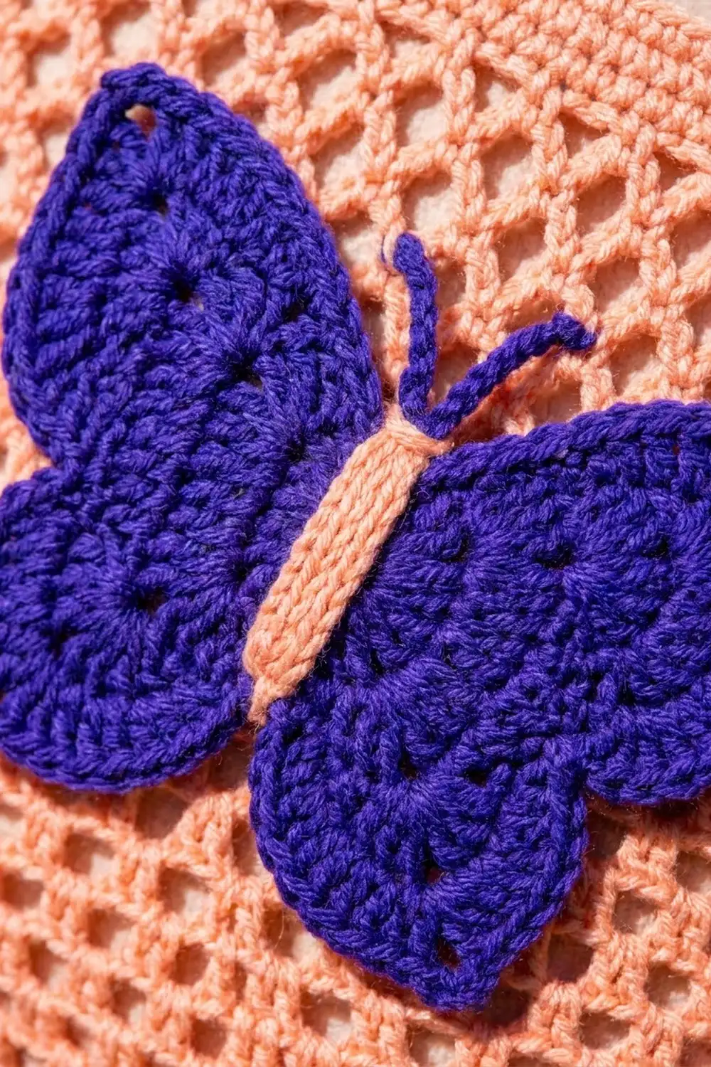 A deep purple crocheted butterfly with peach accents rests on textured coral knit fabric.