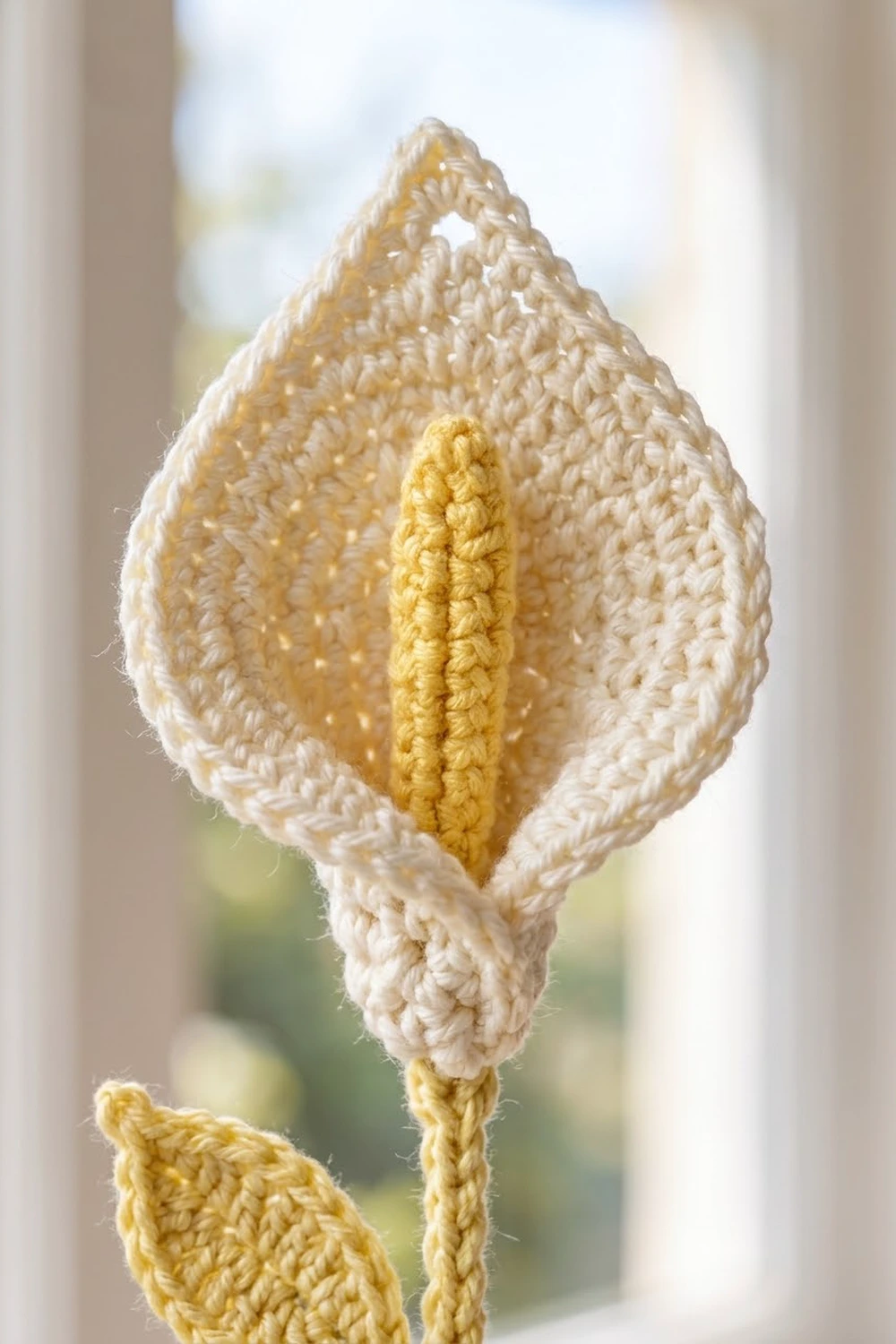 Handmade crocheted flower with cream-white petals and bright yellow center, photographed against soft interior light.