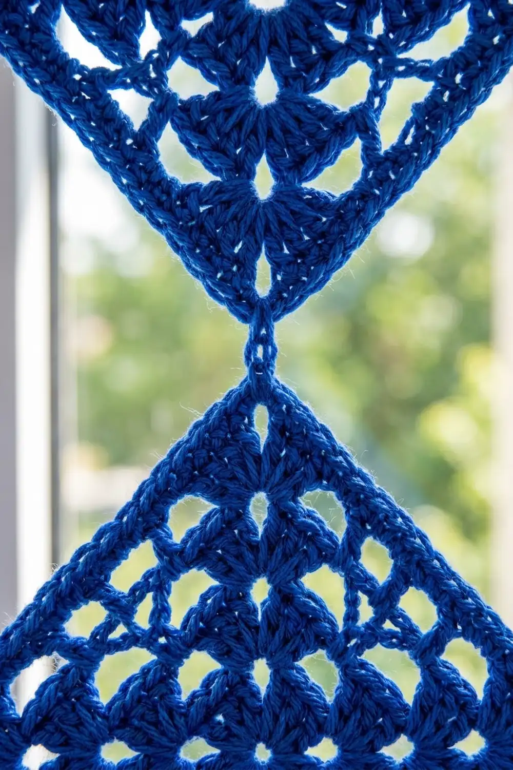A deep blue crocheted textile with intricate open diamond-shaped lacework, held up to a soft, blurred garden background.