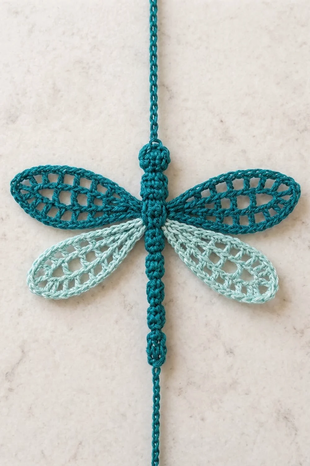 Crocheted teal and pale blue dragonfly ornament with lacy wings hanging against a light marble background.
