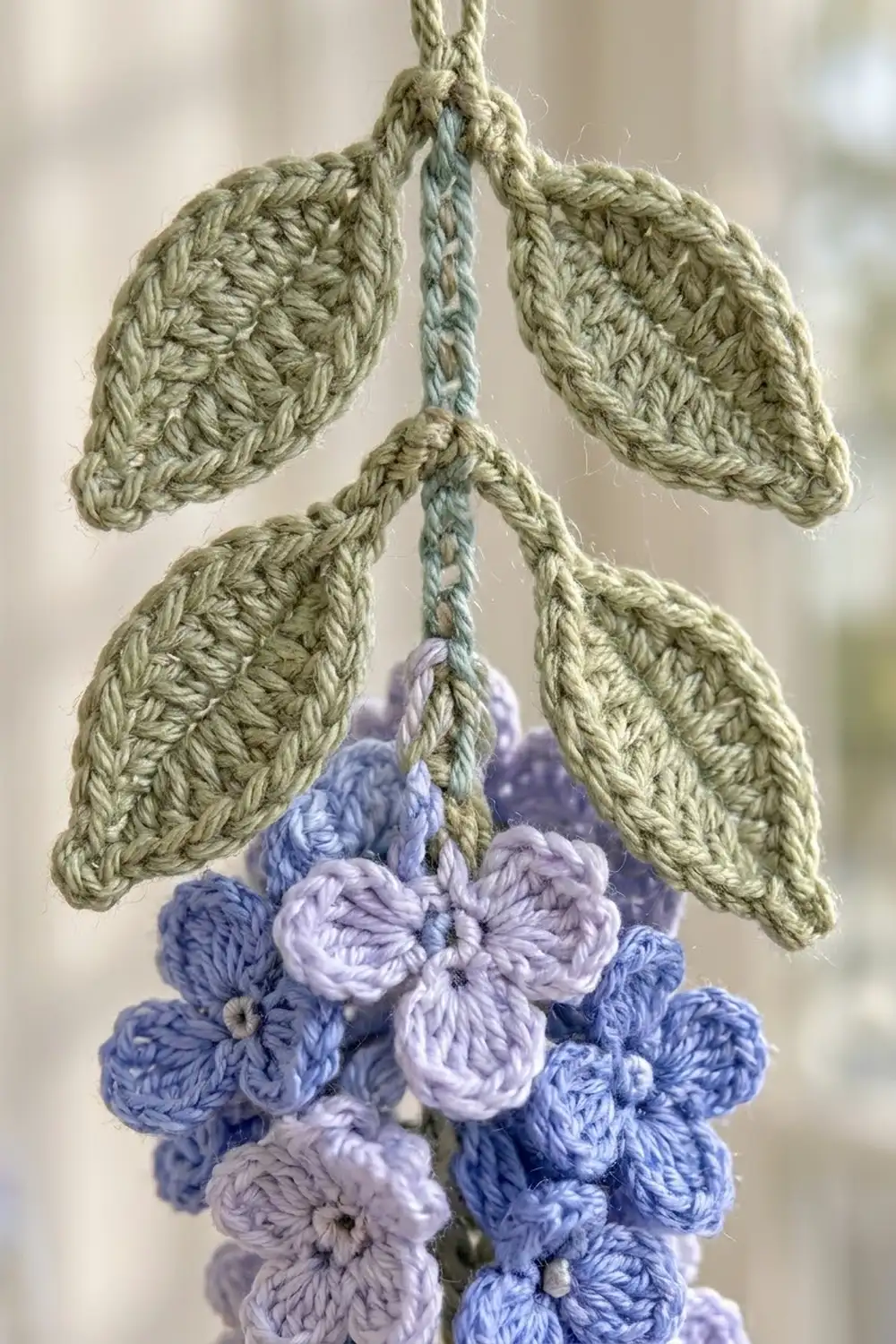 Handmade crocheted plant with sage green leaves and clusters of periwinkle and lavender flowers against a soft, blurred background.
