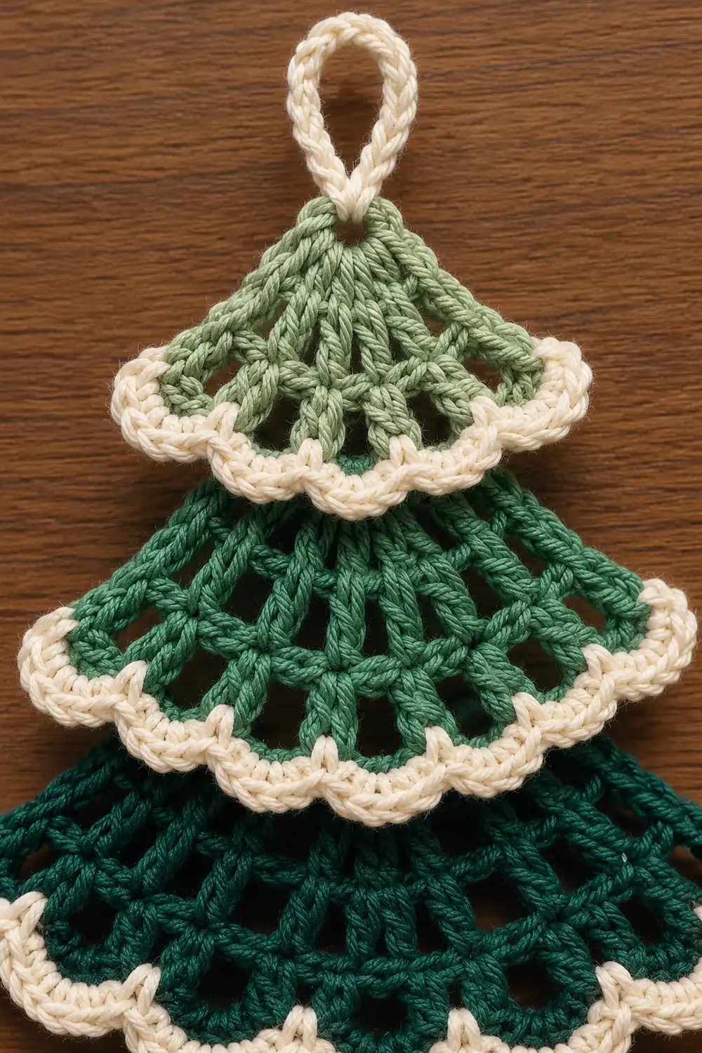 A crocheted Christmas tree ornament with three tiered layers in green and cream colors displayed on wood.
