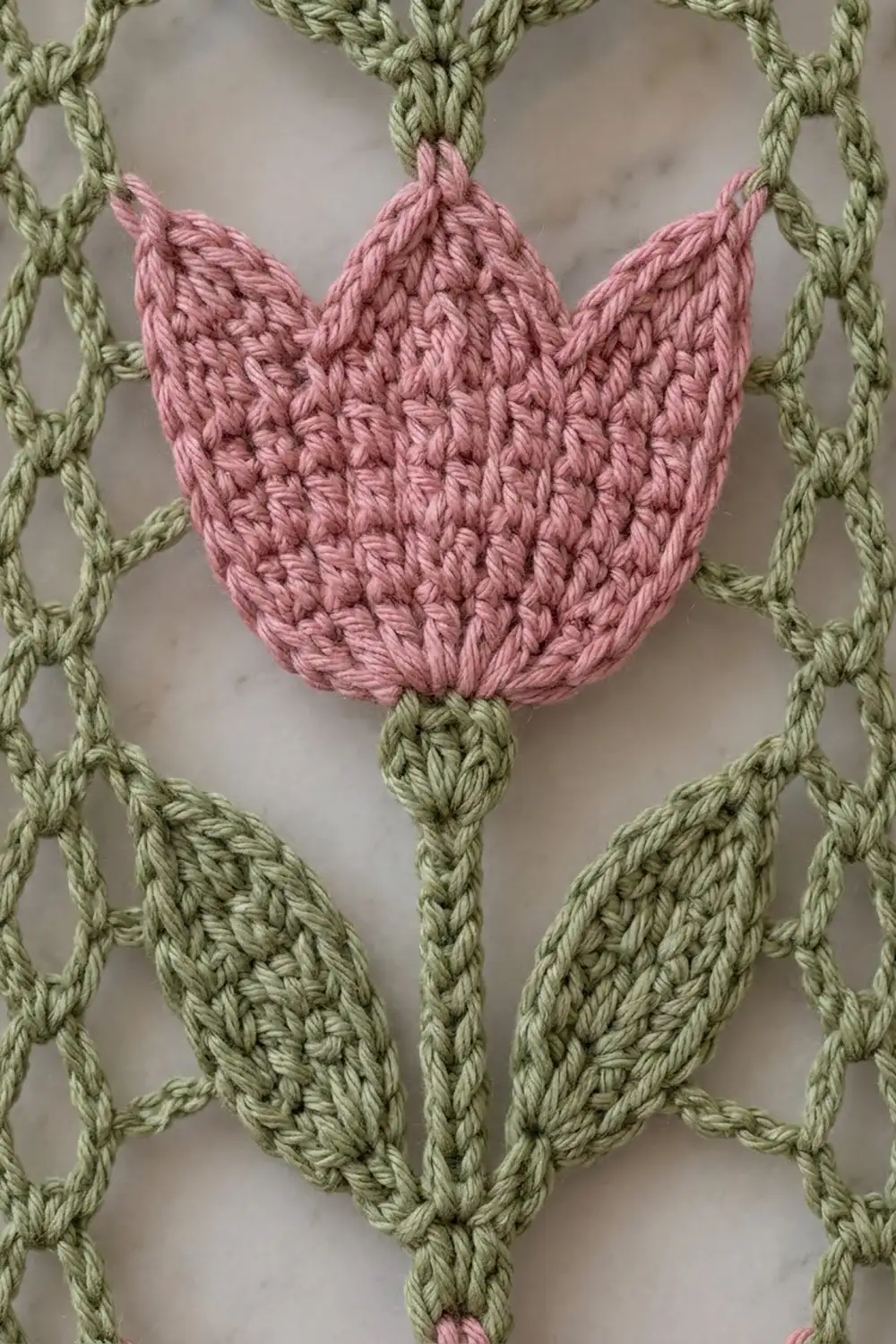 Close-up of a pink crocheted tulip motif surrounded by green twisted cables on a neutral background.