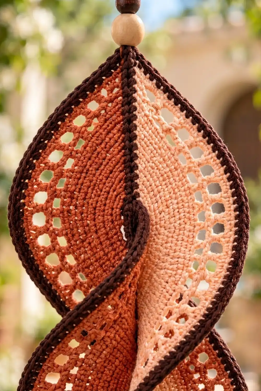 Crocheted teardrop ornament with lacy openwork in rust and peach, topped with wooden beads, displayed outdoors.