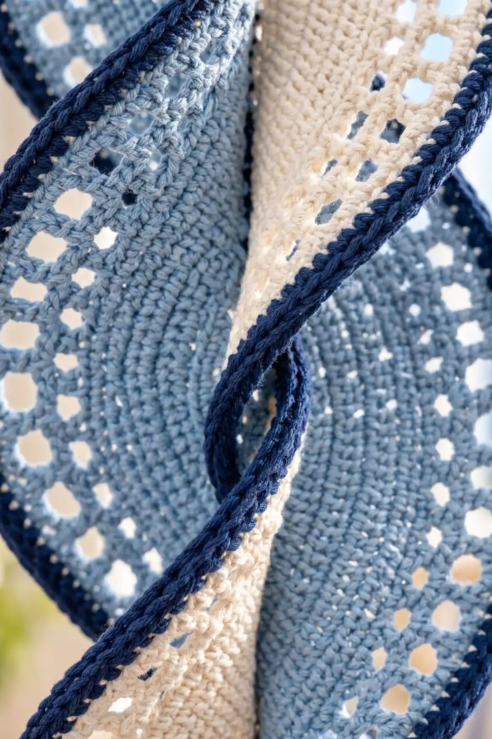 Crocheted fabric in blue-gray and cream with navy binding, showing textured stitches and eyelet holes.
