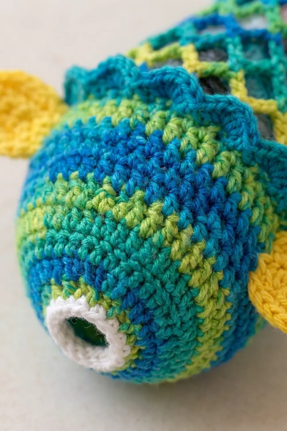 Close-up of striped crocheted toy in blue, teal, lime, and yellow with white eye and visible stitch detail.