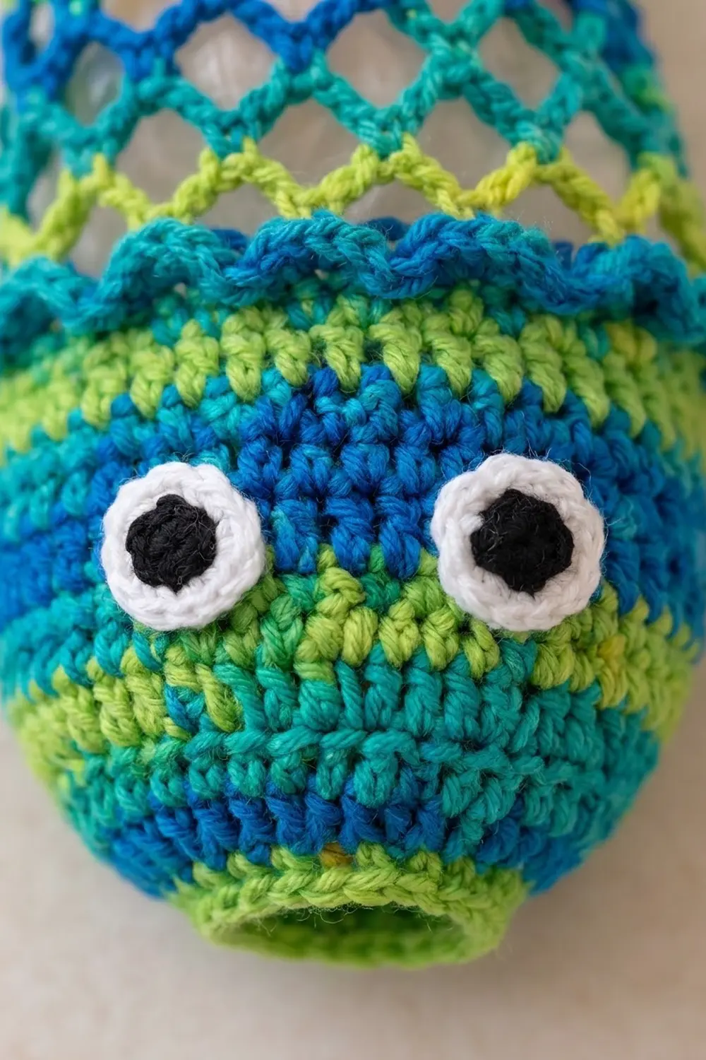 Crocheted monster face with lime-green and blue horizontal stripes, white embroidered eyes, and black pupils.