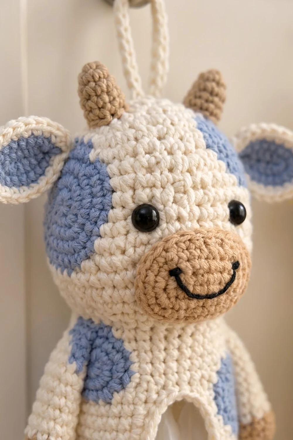 A handmade crocheted cow with cream and blue patches, tan horns, and an embroidered smiling face hanging from white rope.