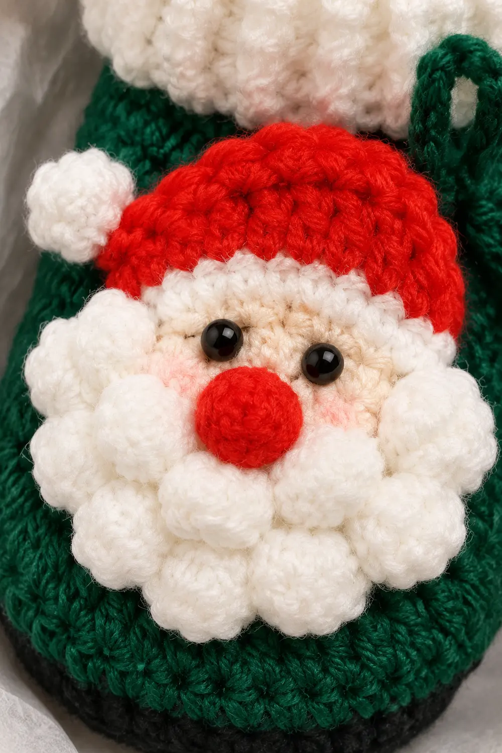 A crocheted Santa ornament with red hat, white beard, and red nose on a green knitted background.