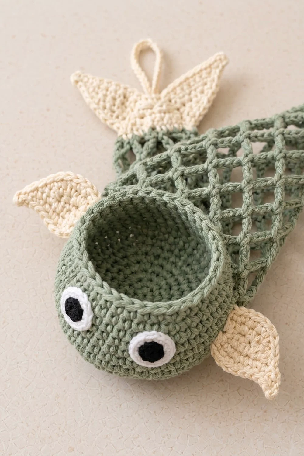 A crocheted sage-green fish with white eyes and cream-colored fins dangling against a soft neutral background.