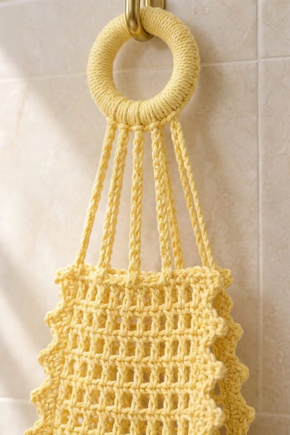 A pale yellow crocheted hanging basket with wrapped rope ring and dangling cord strands against tiled wall.
