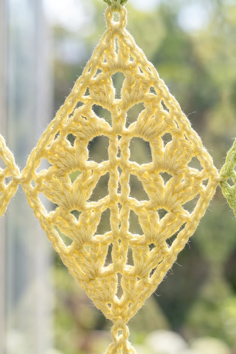 Yellow crocheted diamond pendant with intricate lattice pattern, backlit against soft-focused green foliage.