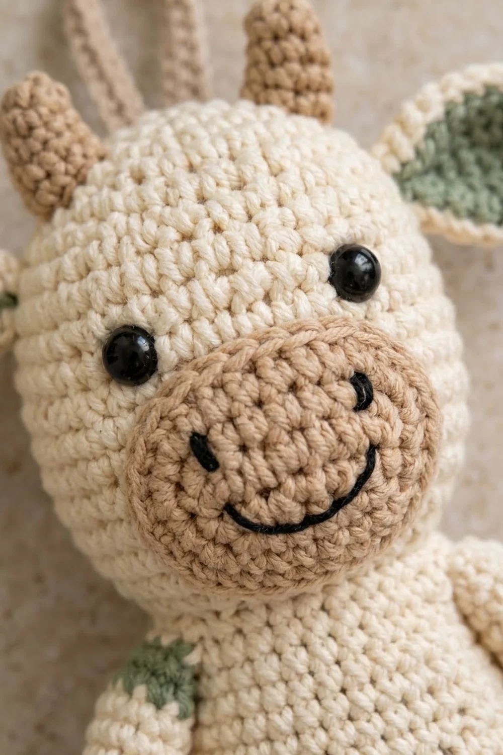 Close-up of a cream and tan crocheted llama amigurumi with black bead eyes, embroidered smile, and blurred green leaves behind.