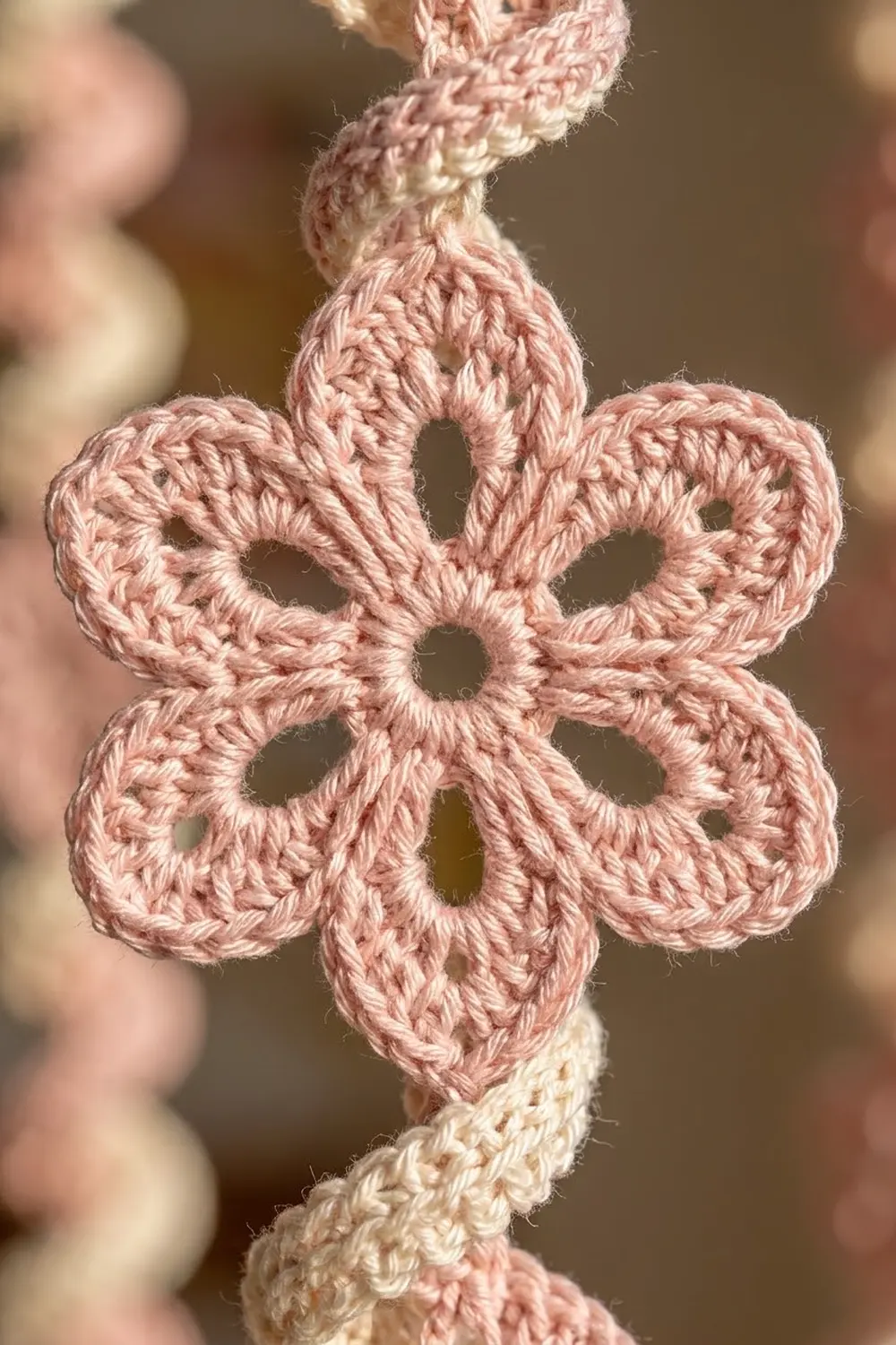 Pink crocheted flower motif with open lacework stitches attached to a cream cord on a soft, blurred neutral background.