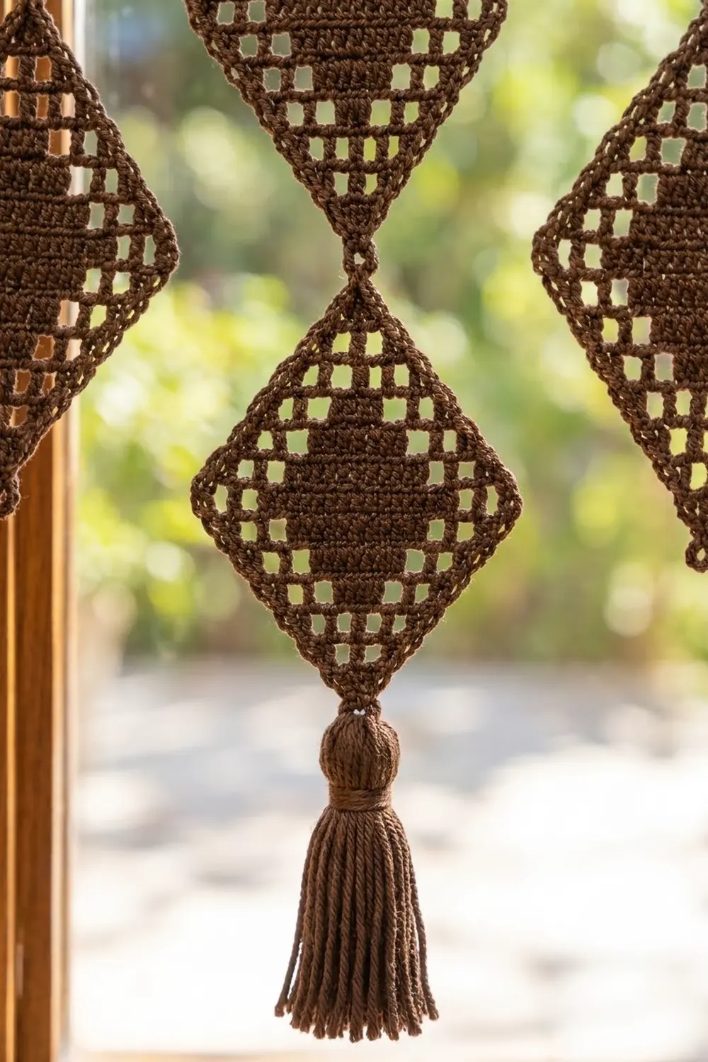 Chocolate-brown crochet ornaments with cream-colored openwork diamond pattern and decorative tassels hanging by a window.