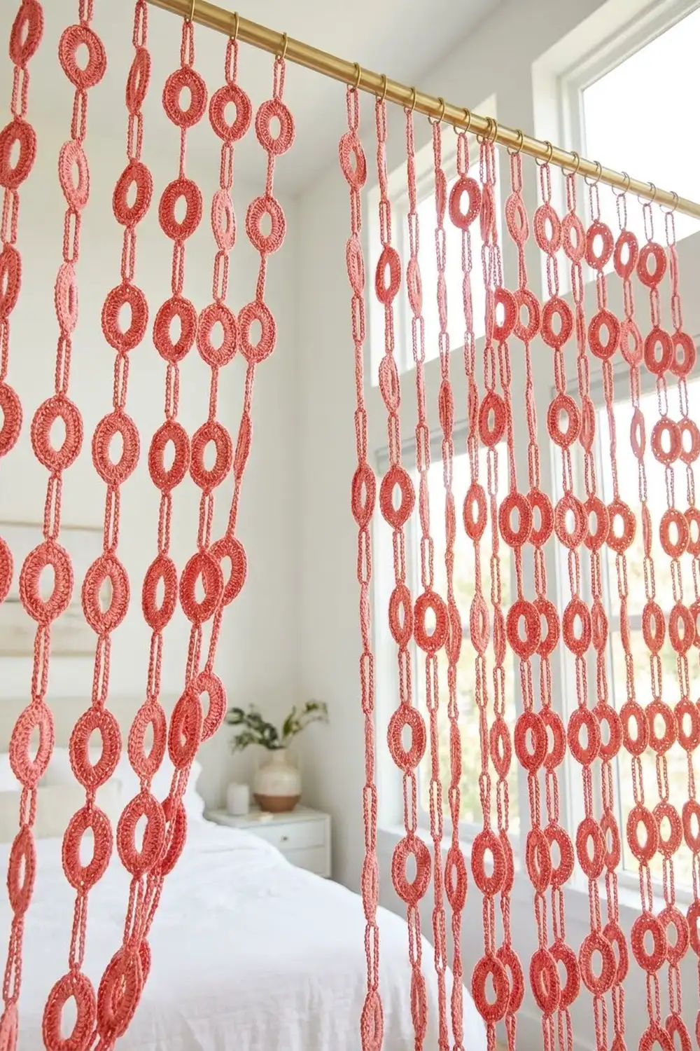 Coral crochet room divider with lacy circles suspended from gold rod above white bed in bedroom.