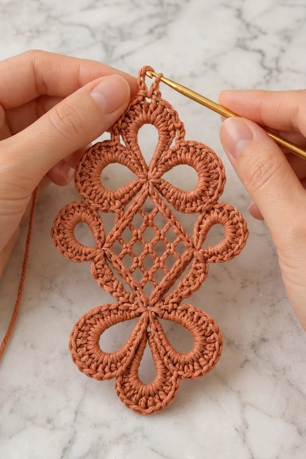 Hands holding a terracotta crocheted lace motif with a gold crochet hook on a white marble surface.