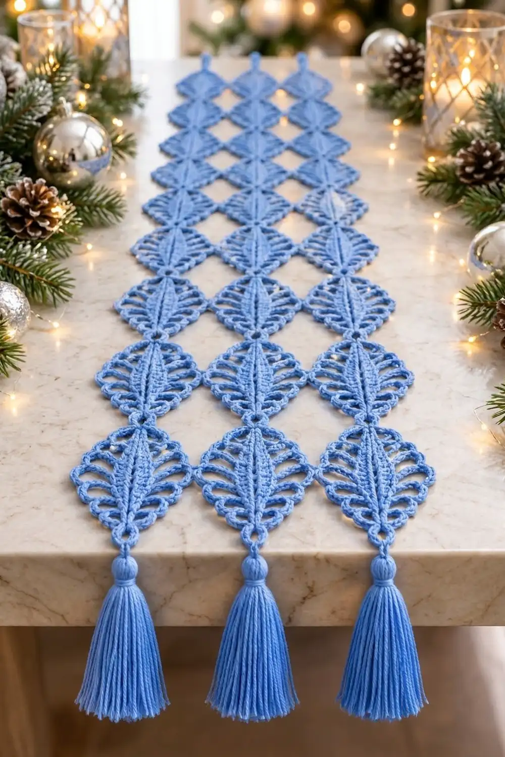 Blue crocheted lace table runner with diamond pattern and three tassels, styled on marble surrounded by holiday greenery and lights.
