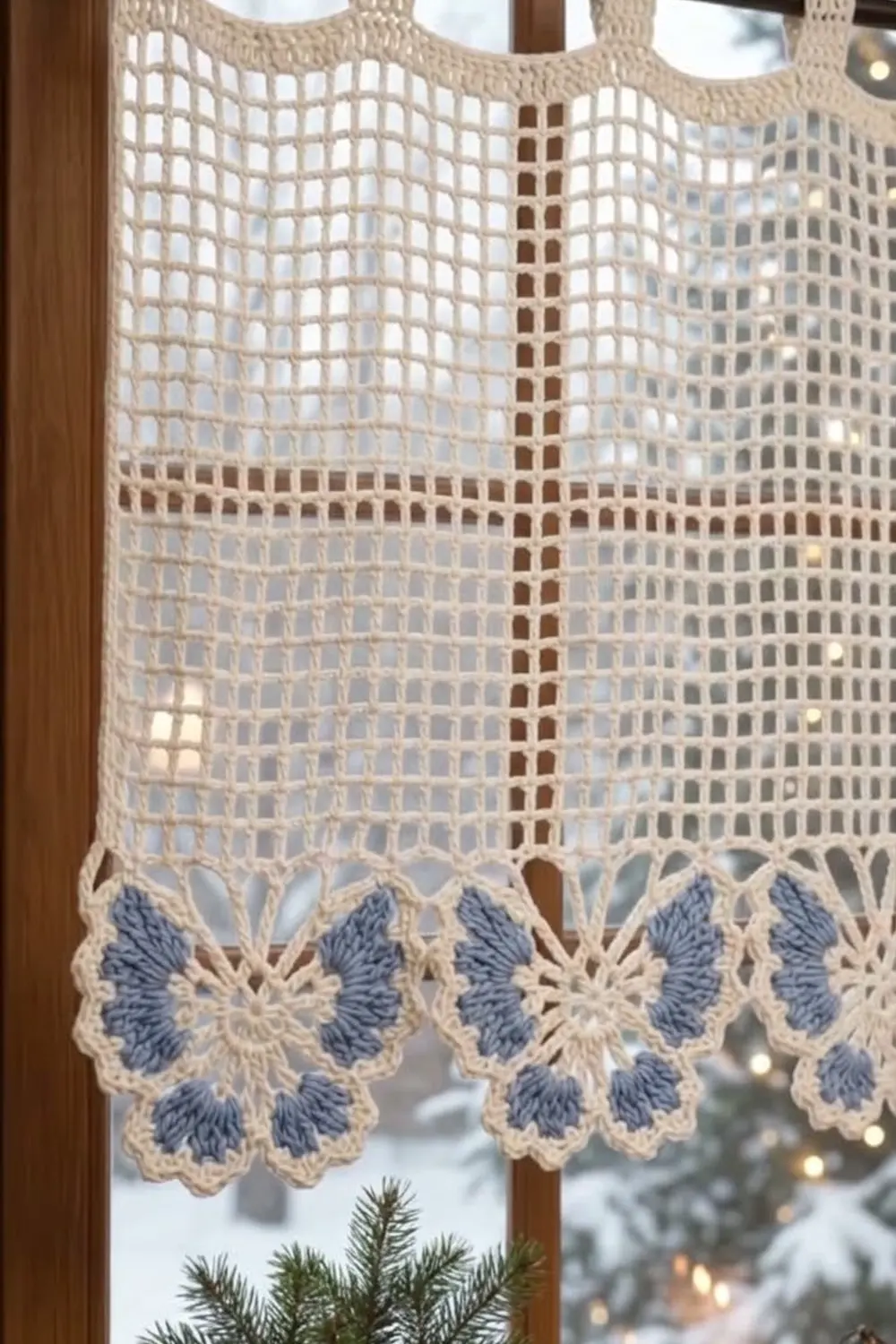 Cream-colored crochet curtain with blue butterfly border hanging in a wooden window frame overlooking a snowy landscape.