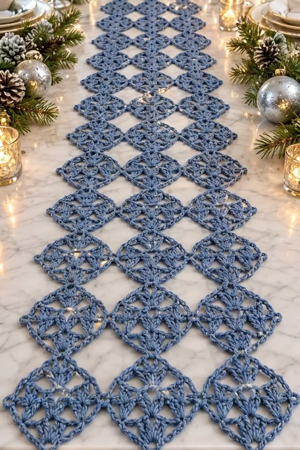 Blue crocheted lace table runner in diamond pattern on marble, styled with evergreen, silver ornaments, and candles for holiday setting.